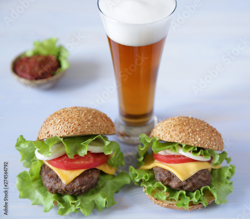 Two hamburger with beef patty, with cheese, pickles, tomatoes, onions, lettuce and tomato sauce in a saucer with a glass of beer on a wooden white background with copy space for text.