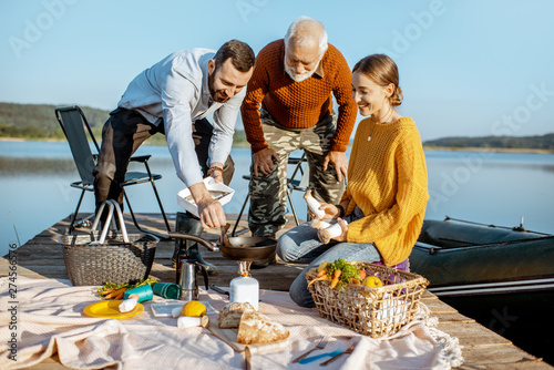 Man and woman with senior grandfather having a picnic with vegetables and fresh caught fish on the lake in the morning