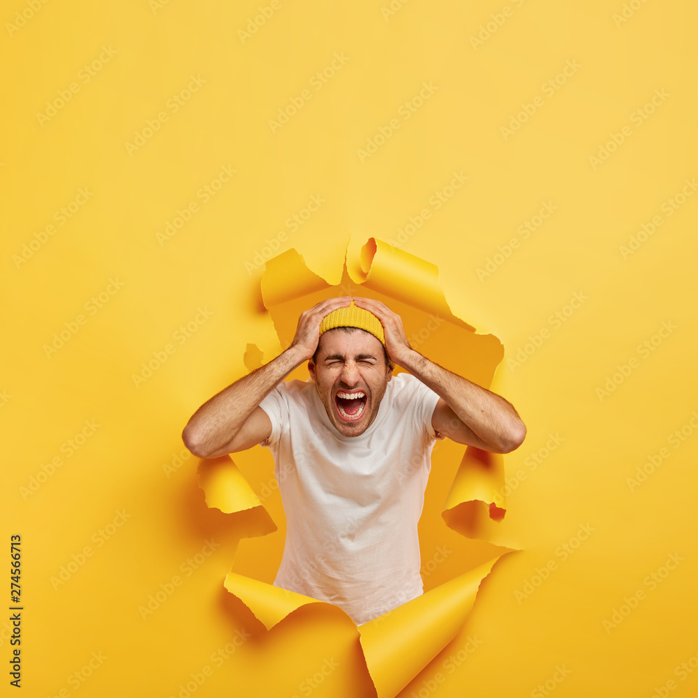 Vertical shot of emotional guy touches head with both hands, wears casual white t shirt, stylish yellow hat, poses in ripped hole paper, shouts from negative emotions, feels annoyed. Bad feelings Vertical shot of emotional guy touches head with both hands, wears casual white t shirt, stylish yellow hat, poses in ripped hole paper, shouts from negative emotions, feels annoyed. Bad feelings