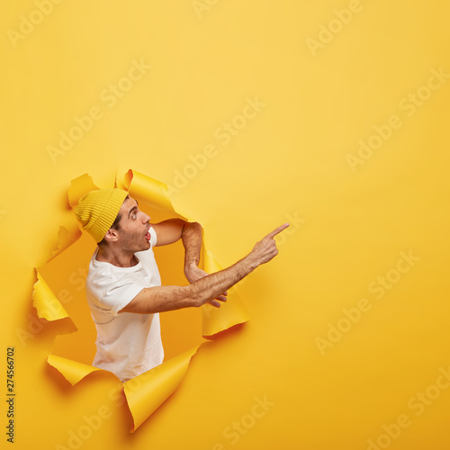 Emotional surprised guy stands in paper hole with ripped yellow edges, demonstrates incredible copy space, says wow awesome, being fascinated and speechless, stands against yellow background