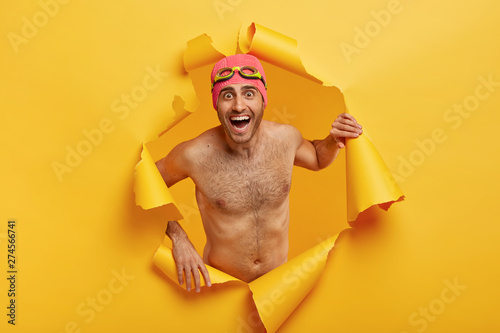 Overemotive man swimmer poses shirtless, wears swimhat and goggles, poses in torn paper hole, laughs happily, notices something awesome, recreats in swim pool, isolated over yellow background photo