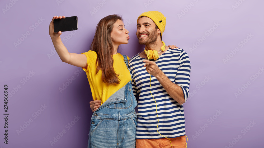 Loving couple stand together, embrace, kiss and take selfie, have perfect relationship. Affectionale girl in yellow t shirt and denim overalls, kisses boyfriend in cheek, holds cell phone, makes photo Loving couple stand together, embrace, kiss and take selfie, have perfect relationship. Affectionale girl in yellow t shirt and denim overalls, kisses boyfriend in cheek, holds cell phone, makes photo