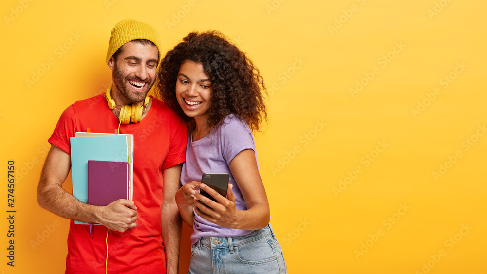 Positive young people focused at smart phone, watch funny video online, dressed in casual clothes, stand closely against yellow background. Happy guy with notepads, looks at girlfriends gadget - obrazy, fototapety, plakaty 