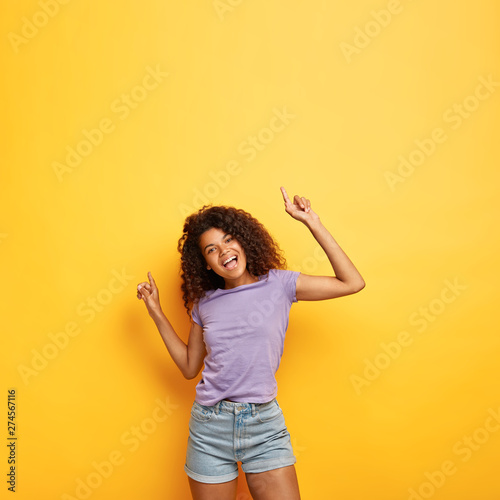 Joyous happy attractive dark skinned female with frizzy hair, raises hands to good vibed music, has slim figure, enjoys party and has fun, dances against yellow background with copy space above