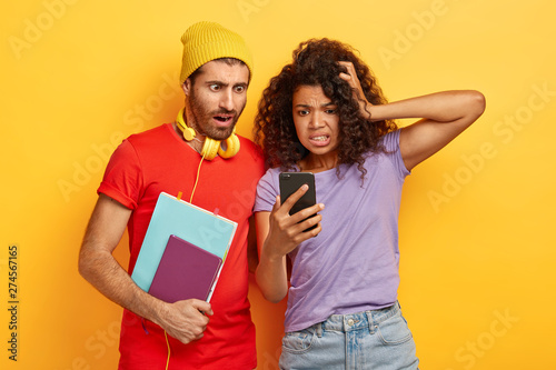 Emotional nervous mixed race students read shocking information from website  stare at smartphone  carry notepad  spend free time during break with technologies. Reaction  techology  studying concept