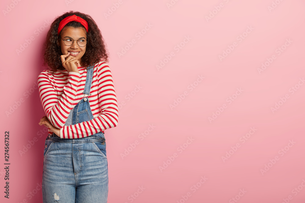 Dreamy positive dark skinned female smiles friendly, listens something  pleasant, has Afro hairstyle, wears casual sweatshirt with red stripes,  denim dungarees, poses over rosy wall, blank space Stock Photo | Adobe Stock