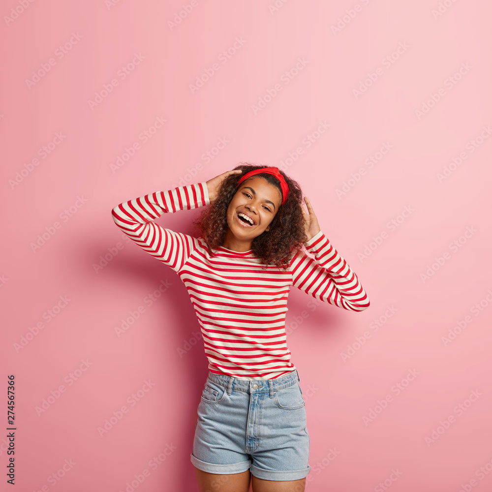 Vertical shot of slim smiling young female model keeps hands behind head,  stands in lazy pose,