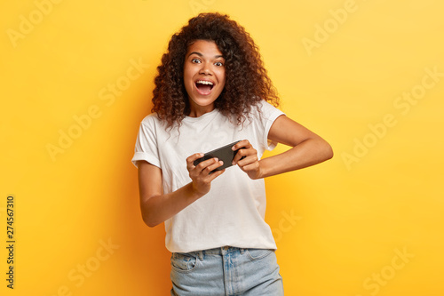 Amused lovely teenager with bushy frizzy hair, makes notes in device, plays online games on cell phone, enjoys leisure time, weekend, online talk in social networks, reads funny article on website