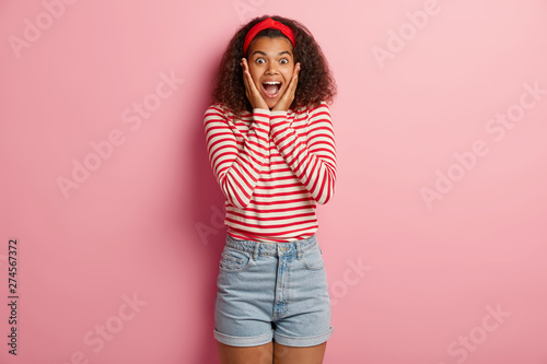 Fascinated happy woman gasps from surprisement, keeps hands on cheeks, attends awesome event, dressed in casual striped jumper, jean shorts, poses against pink background, receives expensive present