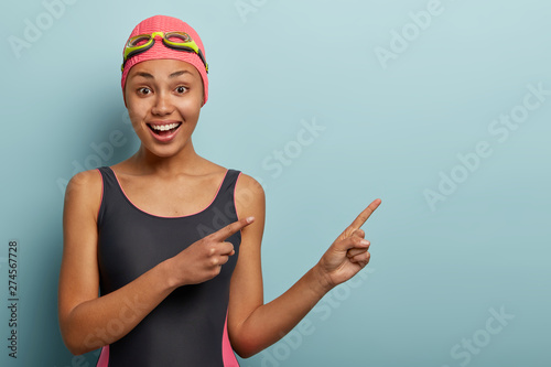 Beautiful happy young woman with charming smile, wears black bikini, swimcap on head, points away with both index finger on copy space, shows direction to beach or swimming pool suggests recreat there