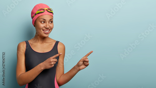 Lovely satisfied female vacationist take break after swimming, points to side, shows free space for your advertising content, dressed in swimsuit and swimhat, spends free time on seaside. Summer promo