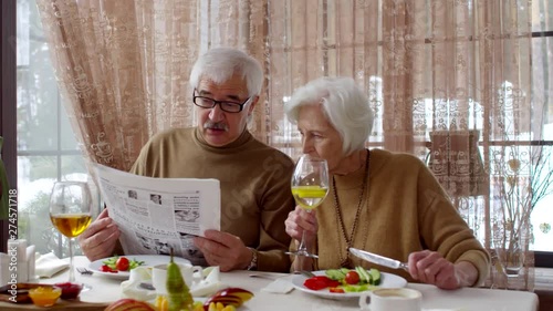 Panning waist-up shot of 60-something Caucasian couple having breakfast together in countryside hotel, man reading newspaper and lady looking at it over his shoulder, discussing news with disapproval photo