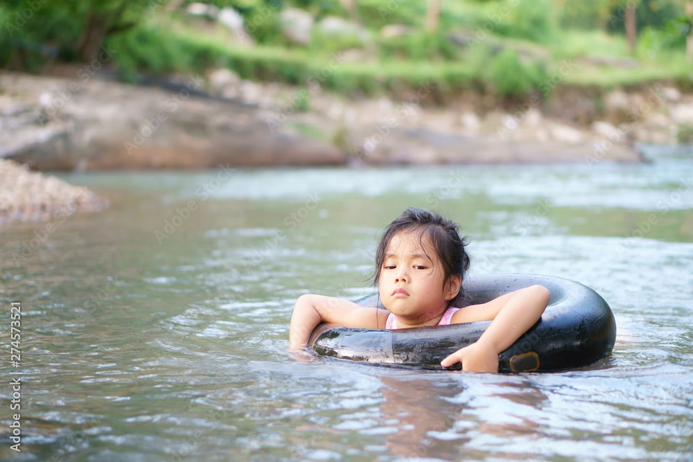 Fototapeta premium Asian child or kid girl with life ring from inner tube or tire for swimming and enjoy playing water with happy on stream or nature river in green jungle or garden forest for summer holiday relax