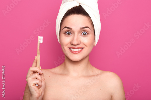 Portrait of smiling pretty woman with astonished facial expression, holds toothbrush isolated over pink background and looking directly at camera, posing with nacked shoulders and white towel on head. photo