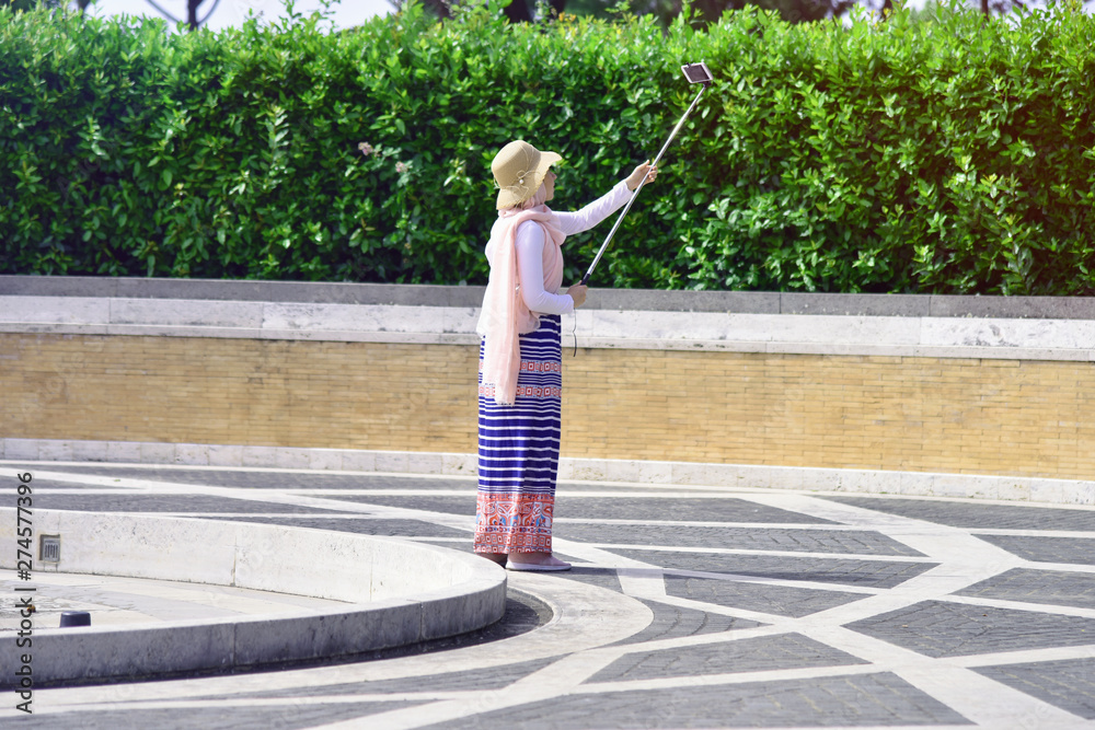 Obraz premium Fashionable beautiful young Muslim European girl in hijab and white shirt taking a selfie on smartphone outdoors in park on sunny day.