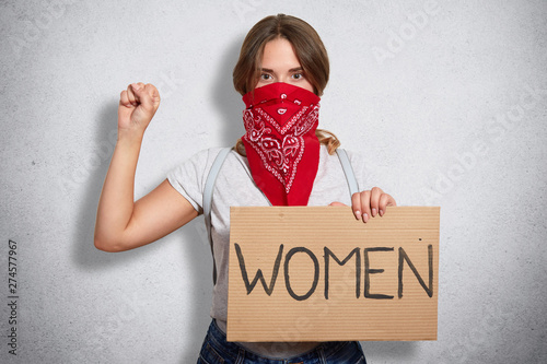 Image of serious bossy militant young feminist raising arm, showing fist, holding sign in one hand, having inscription women on plate, looking directly at camera, fighting for equal women rights.