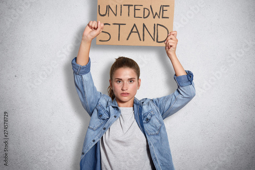 Portrait of militant powerful young feminist showing her fist, holding sign with inscription united we stand in one hand, having decisive facial expression, interested in women social problems. photo