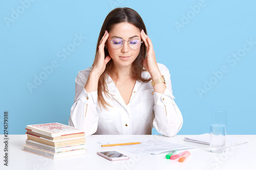 Studio shot of stressed attractive office worker sitting at table isolated over blue background in studio, touching her temples with hands, has terrible headache, looks tired after long hours working. photo