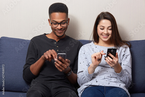 Photo of young woman in casual outfit sitting on couch in room with her African boyfriend and looking at theit smartphones's on smartphone screens and loughing, see something interesting, check email. photo