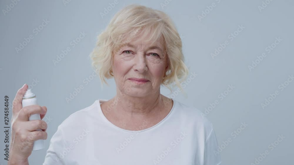 Portrait of cheerful older woman in white shirt with blonde hair against grey background. Senior blonde model sprinkling moisturizing spray on face before makeup. Portrait of cheerful older woman in white shirt with blonde hair against grey background. Senior blonde model sprinkling moisturizing spray on face before makeup.