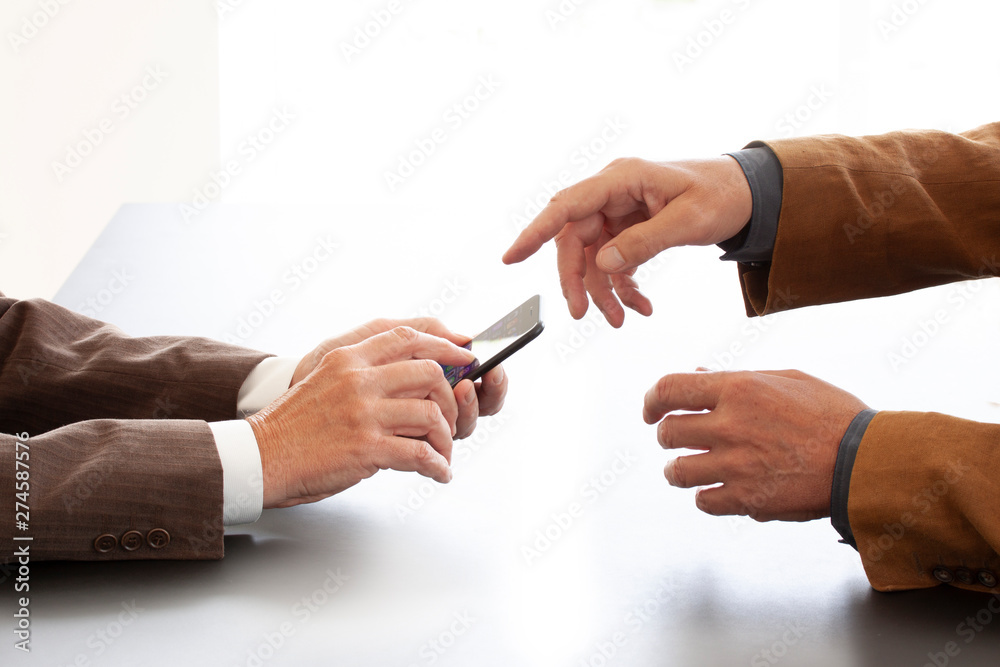 Hands from two businessmen in conversation by a desk. One texting on a cell phone and the other pointing. Negotiating business or a job interview. - Image