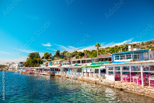 Fototapeta Naklejka Na Ścianę i Meble -  View of fish restaurants or cafes and flowers on beach in Gumusluk, Bodrum city of Turkey. Aegean seaside style colorful chairs, tables and flowers in Bodrum town Marina near beautiful Aegean Sea.