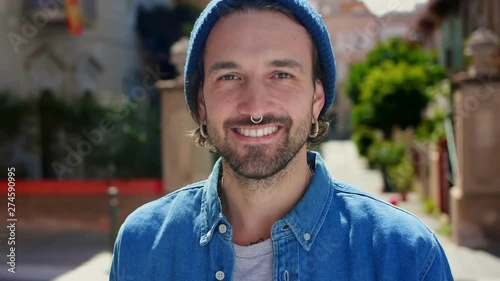 Alluring portrait of smiling trendy man in blue hat standing alone at city landscape, close up of Hispanic confident guy looking to the camera with good emotions, dressed in blue shirt and piercing