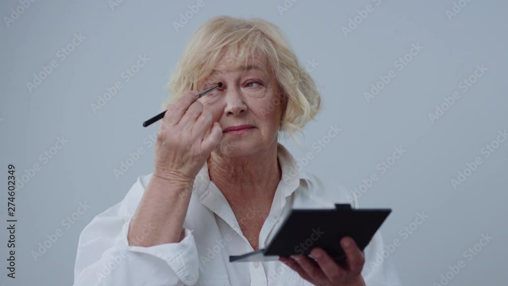 Natural portrait of stunning senior woman with blonde hair and eyeshadow palette applying lovely make-up standing over gray background. Natural portrait of stunning senior woman with blonde hair and eyeshadow palette applying lovely make-up standing over gray background.