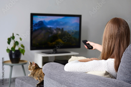 A young woman watching TV while sitting with her cat on sofa in living room at home. Nature, documentary, tv screen, binge watching