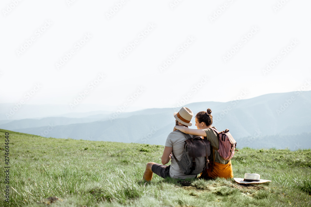 Young couple sitting on the green meadow, traveling with backpacks in the mountains during the summer time