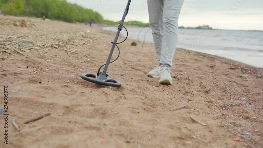 Tracking left closeup of unrecognizable woman with metal detector searching for valuable objects walking on beach along sea coastline