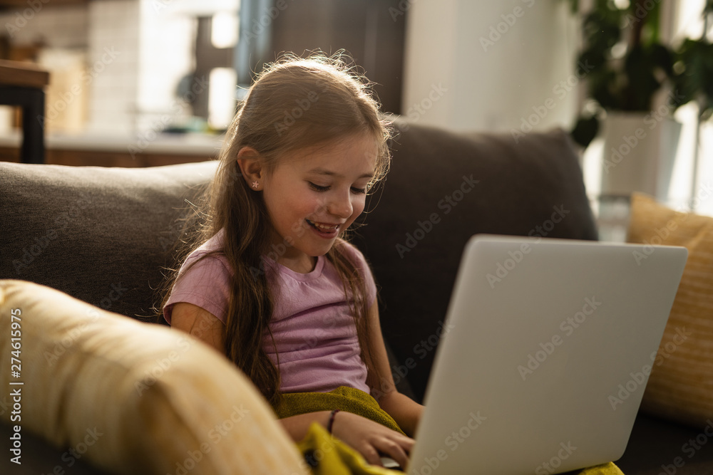 Naklejka premium Smiling little girl using laptop on the sofa at home.