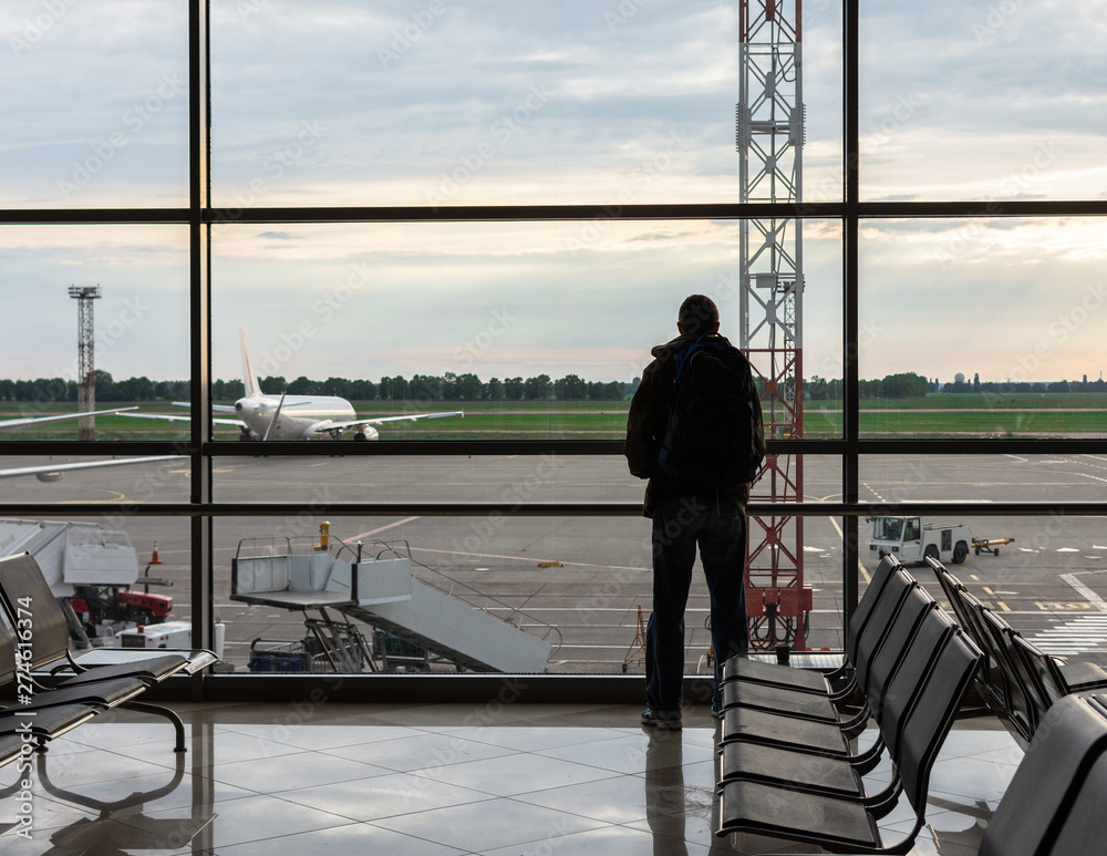 Man with a backpack on his shoulders looks out the window at the planes at the airport, air tourism Man with a backpack on his shoulders looks out the window at the planes at the airport, air tourism