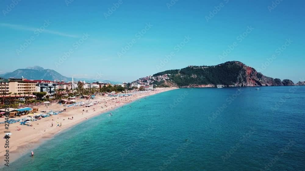 Aerial view: Cleopatra beach and azure sea, in the background a mountain and a fortress in the Turkish city of Alanya. Many people swim and sunbathe on the beach. Sunny, clear day, blue sky. Aerial view: Cleopatra beach and azure sea, in the background a mountain and a fortress in the Turkish city of Alanya. Many people swim and sunbathe on the beach. Sunny, clear day, blue sky.