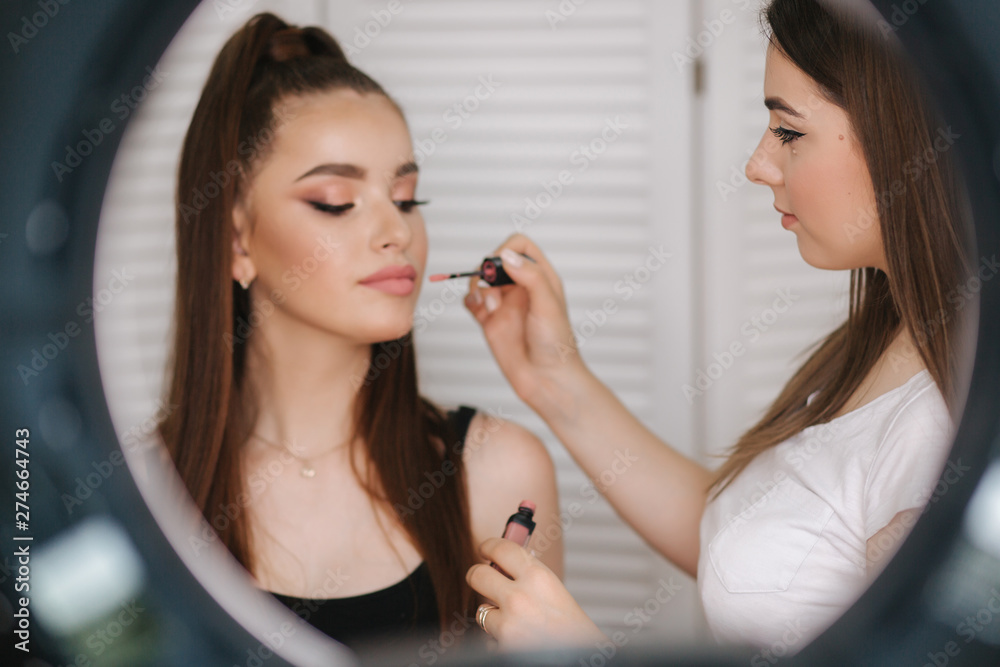 Photo though the ring lamp of makeup artist doing a makeup for attractive young girl. Background of white folding screen