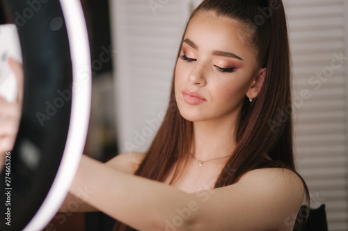 Young attractive model make a selfie after makeup in beauty studio. She use a ring lapm for photo. Background of white folding screen photo