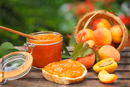 Apricot jam in a glass jar, fresh apricots in a basket on wooden background. Sweet breakfast in nature.