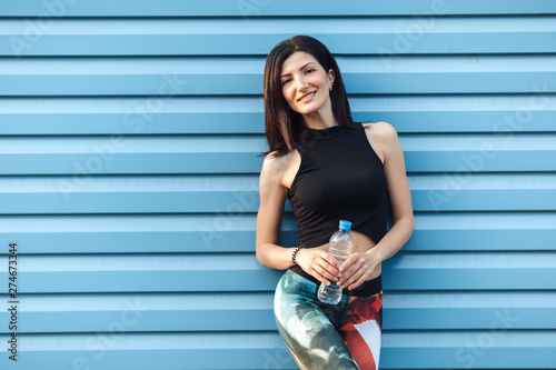 Beautiful young fitness brunette girl in the top and the leggings with water bottle standing near blue wall looks at the camera and smiles. © lena_itzy