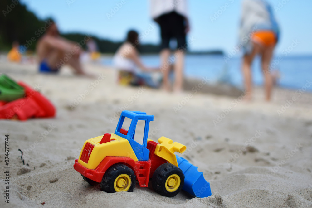 Toy tractor bulldozer on beach sand and blurred silhouette of people and sea on background. Leisure time with friends and family and summertime vacation concept. Toy tractor bulldozer on beach sand and blurred silhouette of people and sea on background. Leisure time with friends and family and summertime vacation concept.