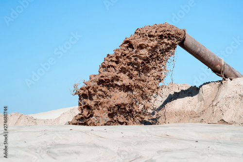 The flow of brown liquid from the pipe on the background of a pile of sand and blue clear sky. photo