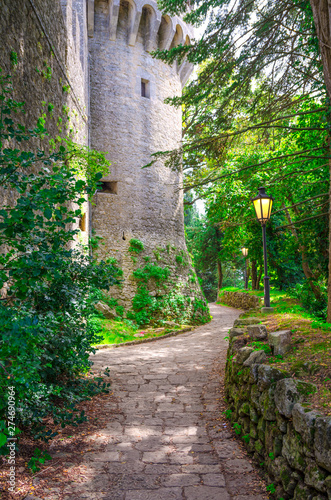 Fototapeta Naklejka Na Ścianę i Meble -  Cobblestone path with street light lamp near wall of stone brick medieval castle tower in green park forest in Republic San Marino