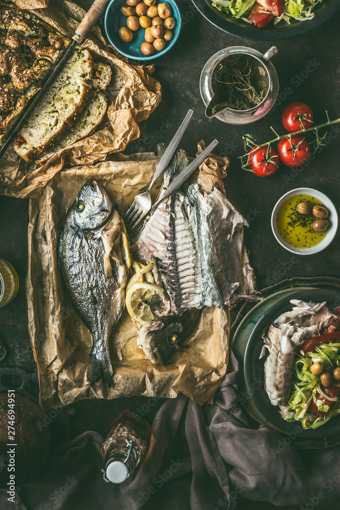 Fototapeta premium Dinner table with backed dorado fish, salad and homemade bread on dark rustic table, top view. Mediterranean lunch or dinner. Healthy food