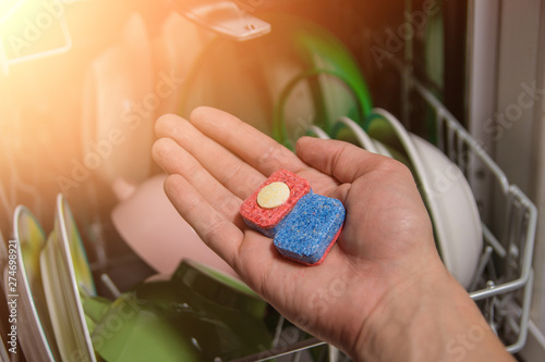 dishwasher tablet. An open dishwasher with clean dishes in a white kitchen, front view. dishes, selective approach. white dishes. colored dishes. spoons forks. hand with a pill. chemical agent