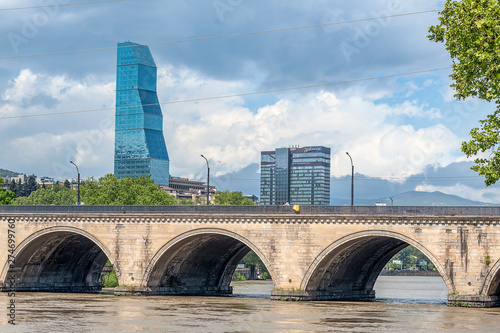 Georgia, Tbilisi, view of the glass tower of the Biltmore Hotel in the center of the city against the background of a blue sky and an ancient stone bridge in the foreground photo
