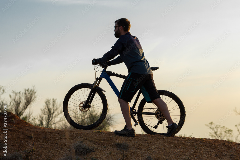 Fototapeta premium Cyclist in shorts and jersey on a modern carbon hardtail bike with an air suspension fork rides off-road on the orange-red hills at sunset evening in summer 