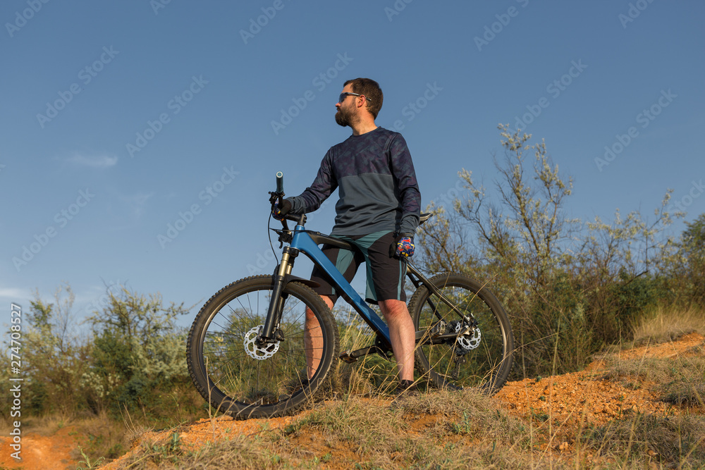Cyclist in shorts and jersey on a modern carbon hardtail bike with an air suspension fork rides off-road on the orange-red hills at sunset evening in summer Cyclist in shorts and jersey on a modern carbon hardtail bike with an air suspension fork rides off-road on the orange-red hills at sunset evening in summer