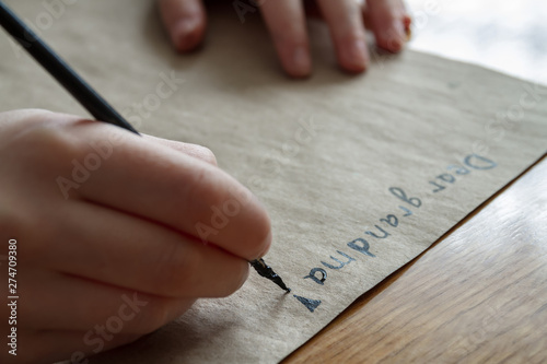 A small girl writes a letter to dear grandma by black paint on brown paper. Closeup, selective focus