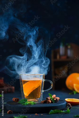 Double wall glass cup with citrus tea, cinnamon, and mint leaves. Steam in dark food photography with copy space.