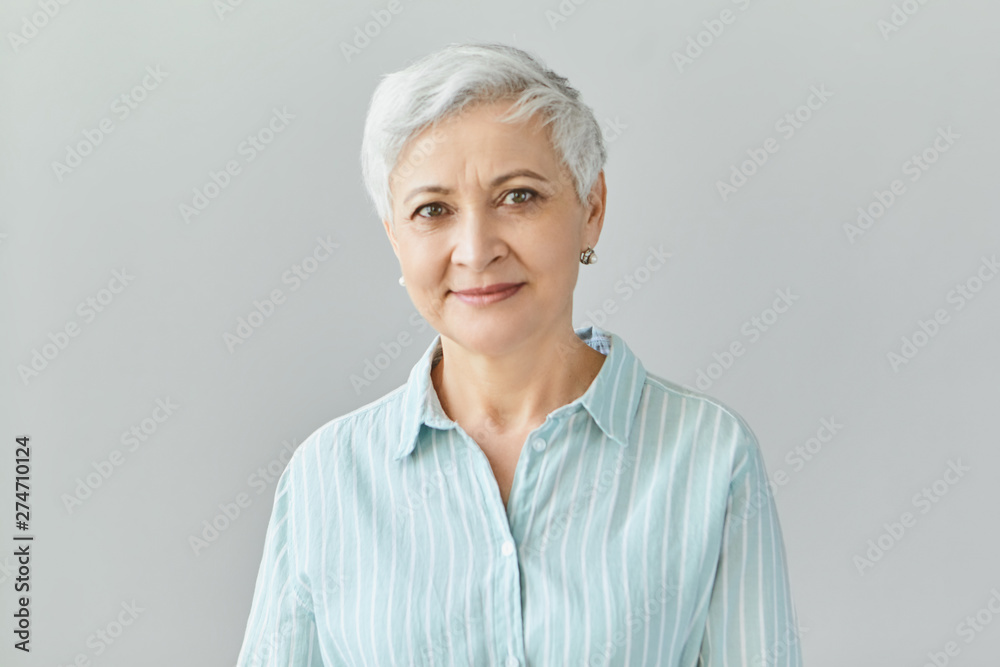 Isolated image of friendly good looking European businesswoman with pixie gray hair smiling confidently at camera, glad with work results of her team, posing in studio, dressed in formal striped shirt - obrazy, fototapety, plakaty 