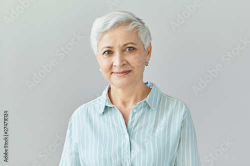Isolated image of friendly good looking European businesswoman with pixie gray hair smiling confidently at camera, glad with work results of her team, posing in studio, dressed in formal striped shirt